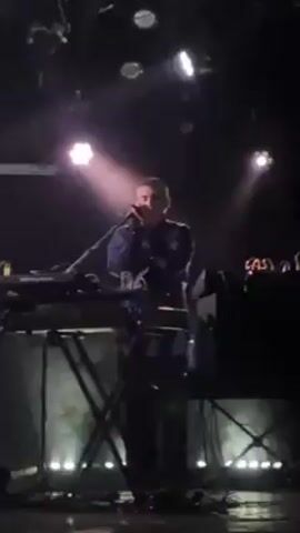 Here's a detailed description of the video:

A musician is performing live on stage. The person, a fair-skinned individual, is wearing a dark blue button-down shirt and dark pants. The musician is positioned behind a keyboard and other electronic music equipment, which sits on a silver metal stand. A microphone is in front of the musician, who is singing into it. Two bright spotlights are positioned above and behind, casting light on the performer and creating a somewhat dramatic effect. Additional stage lighting is visible on the ceiling. Cables and wires sprawl across the stage, connected to the musical equipment. The background is dark, suggesting an indoor concert venue. The image is slightly shaky, indicating it was likely filmed with a handheld device.