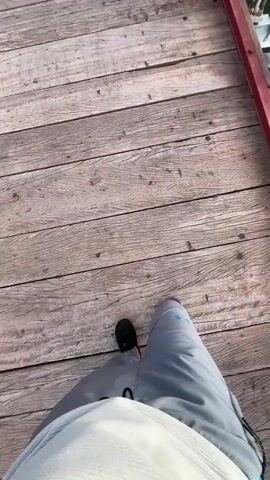 The video starts with a view of a person's feet, wearing brown shoes and light grey pants, walking on a wooden plank bridge. The camera then moves to show the side of the bridge, with a red railing, the water below, and another bridge above. The second bridge is concrete and supported by several pillars in the water. The sun shines through the second bridge, reflecting on the water below. The sky is visible with patchy clouds.