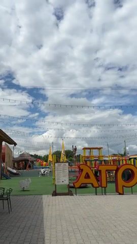A vertical shot shows an outdoor scene with a large sign in the foreground that reads "АГРОЛЕНД" in red and yellow letters. To the right of the sign is a large stuffed animal resembling a tiger or a cow with yellow and red stripes. In the background, there is a playground with colorful structures, inflatable attractions, and people walking around. String lights are strung across the sky. The sky is cloudy with patches of blue. The ground is paved with interlocking stones. Buildings with red roofs are visible in the distance.