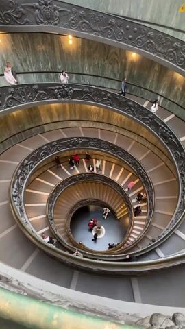 The video shows a downward aerial view of a grand, ornate, spiral staircase within a building. Many people are seen ascending and descending the staircase. The staircase has gray stone steps and dark metal railings decorated with intricate designs. The walls are clad in a light green marbled material. The lighting is provided by warm-toned lamps. The bottom of the staircase features a central feature, possibly a fountain or sculpture, where several people are gathered.