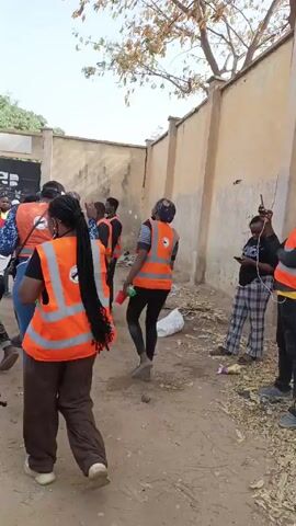 The video shows a group of people in an outdoor area with a wall on one side. Most of the people are wearing orange safety vests, and some are holding or carrying small bottles or containers. Some of the people are looking toward a large gate in the background. Trash and debris are scattered on the ground. Some of the people are talking and gesturing. A silver car is parked near the group.