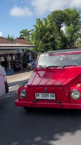 The video shows a red, vintage Volkswagen Thing convertible driving down a street. The car has a black soft top that is partially down. The license plate reads "DK 1659 QQ" and appears to be from Indonesia. The car is a bright red color and looks to be in good condition. The car is moving from left to right in the frame. The background shows a street with some buildings and trees. The sky is blue and sunny. The video is shot from a stationary position on the side of the road.