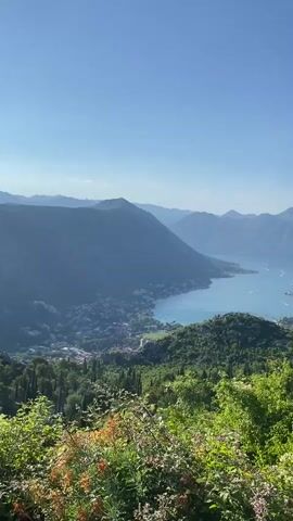The video pans across a scenic vista of a bay surrounded by mountains. Lush green trees and foliage are in the foreground, with a stone wall visible on the right side of the frame. In the middle ground, a town or city is nestled along the shoreline, with buildings clustered together. The bay itself is a calm body of blue water, with a few boats dotting the surface. The background is dominated by a series of majestic mountains, their peaks shrouded in a light haze. The sky is a clear, bright blue, indicating a sunny day. The camera movement is slow and deliberate, allowing the viewer to take in the expansive landscape.