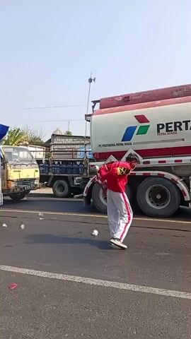The video shows a roadside scene with several vehicles and people. A large Pertamina fuel tanker truck is prominent, with its logo and warning symbols visible. To the left of the truck, there's a yellow truck. In the foreground, several individuals are present. One person in a red jacket and white pants with red stripes is holding what appears to be a baton or flag pole. Another person in a black t-shirt and a sarong is holding a cardboard box. A woman in a hijab is seated on a motorcycle. The road surface is visible with some debris scattered on it. The environment appears to be outdoors with a clear sky. The people seem to be interacting or moving in the vicinity of the vehicles.