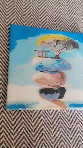 This is a medium shot of a painting placed on a chevron-patterned rug. The painting depicts a surreal landscape with a stack of smooth, colorful rocks, balanced precariously. Atop the highest rock, a solitary, whimsical tree with bright yellow foliage stands, beside a small, red torii gate. The background is a sky of soft blues and whites, with wispy clouds, suggesting a serene and dreamlike atmosphere. The horizon line is low, with hints of a pale blue and white body of water or mist below the rocks. The painting's style is impressionistic, with visible brushstrokes and a gentle blend of colors. No people are visible in the painting or the surrounding environment. The lighting suggests daytime, possibly late afternoon, due to the soft, diffused light. There is no visible text in the media file. The setting is indoors, with the painting resting on a rug, and the background suggests a soft, neutral tone. The overall mood is peaceful and contemplative, evoking a sense of balance and tranquility.