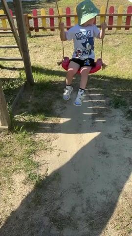 A young child wearing a Sonic the Hedgehog t-shirt, shorts, a green bucket hat, white socks with cartoon characters, and white and blue sandals sits on a red swing. The child is holding the swing ropes and is looking to the right. The child is on a playground with a wooden swing set structure. In the background, there is a colorful fence and grass. The ground around the swing is sandy and covered in shadows. The video shows the child on the swing.