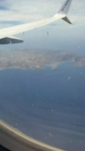 From an airplane window, a view of the ocean and sky is seen through a dirty window. The airplane’s wing is visible. Two islands are visible far below in the blue water.