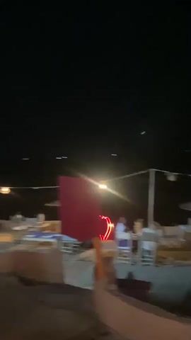The video captures a nighttime scene at an outdoor establishment, likely a bar or restaurant, in Douar Ait Daoud, Morocco. The setting appears rustic with a thatched roof structure and natural materials. There are several people visible, engaged in various activities.

In the initial frames, the scene is blurry, suggesting movement. A stage or elevated area with bright, patterned lighting is visible, and individuals are on or near it. Further into the video, the focus shifts to a bar area. Two men are behind the counter, with one appearing to be serving or preparing drinks. Another man, partially obscured, is seated closer to the camera. The lighting is a mix of warm incandescent and vibrant blue, creating an atmospheric ambiance. Decorative lanterns hang from the ceiling.

The time of day is clearly nighttime, indicated by the darkness of the sky and the artificial lighting. There are no strong weather cues, but the outdoor setting suggests a pleasant evening. The overall mood seems lively and social, consistent with a gathering at a bar or restaurant. There is a visible "Coca-Cola" logo on a refrigerator unit behind the bar. The activity appears to be a social gathering, possibly with entertainment, given the stage-like structure.
