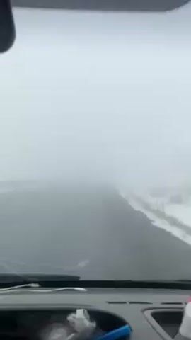 The video shows the view from inside a car driving on a road with heavy fog. The windshield wipers are on. The road is gray and the guardrail is visible on the left side of the road. There appears to be snow on the side of the road. The dashboard is visible with some items including crumpled paper and a charging cable.