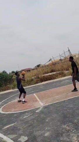 The video depicts two men playing basketball on an outdoor court under a cloudy sky. The court is surrounded by a fence and tall grass. In the initial frame, one man in a black shirt and grey shorts is shooting the ball towards the hoop. He is standing on the right side of the court. Another man is standing to his left, partially out of frame. As the video progresses, the shooter catches the rebound and dribbles the ball to the opposite side of the court, preparing for another shot from behind the three-point line. The basketball hoop is black and white, with a simple design. Buildings can be seen in the background.