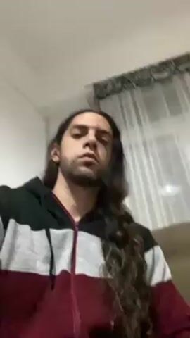 The video shows a man with long, curly brown hair wearing a hoodie, sitting on a brown couch. He is indoors with a white wall behind him. There is a curtain to his left and a plant on the wall behind the couch. The man raises a light green can and drinks from it, looking into the camera.