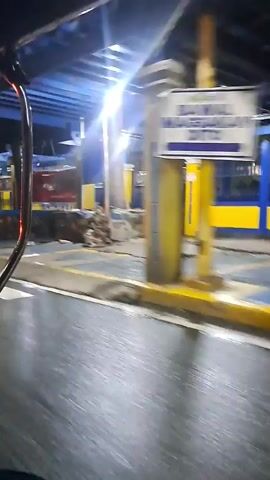 The video shows a street-level view of a covered area, possibly an establishment or a bus terminal, at night in Mandaluyong, Philippines. The scene is illuminated by bright overhead lights, casting strong highlights and shadows. We see a paved road with white and yellow painted lines, suggesting a pedestrian crossing or road markings. The building features a blue ceiling and a series of yellow pillars supporting the roof. The walls are constructed with a mix of stone and blue paneling. 

There are glimpses of people in the background, one of whom appears to be sitting or resting on a low wall. Signs are visible on the walls, but the text is too blurry to read. The overall impression is of a busy, well-lit public space at night, with ongoing activity despite the darkness. The wet appearance of the road might indicate recent rain or a cleaning effort. The dynamic motion of the camera suggests the footage was captured from a moving vehicle.