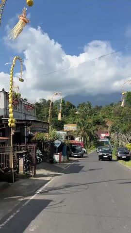 The video shows a street scene with a road, several vehicles, and buildings. On the left side of the road, there are parked motorcycles, a building with a sign, and a small stand with blue tarp. A tall, decorative archway is also visible on the left. The road ahead curves to the right and is lined with trees and vegetation on both sides. Several cars and a truck are driving on the road. In the background, there are hills or mountains covered in greenery and a blue sky with white clouds. The time of day appears to be daytime, likely late morning or early afternoon, given the lighting.