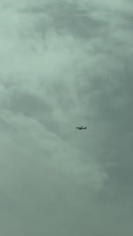 The video shows a passenger airplane flying through a cloudy sky. The plane is small in the frame and appears to be moving from left to right. The clouds are dense and gray, obscuring the sun and creating a muted atmosphere. The aircraft has a distinctive red and white livery. The footage is steady and focused on the airplane and its surroundings.