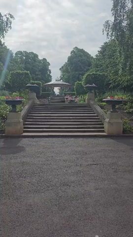 The video shows a wide outdoor shot of a park with a paved walkway in the foreground leading to a set of stone stairs. The stairs are flanked by large, decorative urns filled with flowers. At the top of the stairs, there is a gazebo with a red roof, surrounded by trees and flower beds. The sky is overcast, and the trees are lush and green. The video pans slowly up the stairs towards the gazebo, giving the viewer a sense of walking through the park. There are no people or animals visible in the video. The overall impression is one of a peaceful and well-maintained public space.