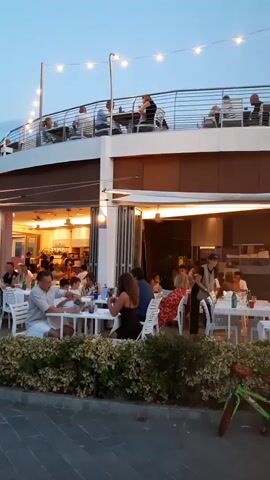 The video shows an outdoor dining area with several tables set up on a paved area. People are seated at the tables, eating and talking. Some tables are occupied by families, while others are occupied by couples. There are also some people standing and walking around. In the background, there is a building with a balcony where more people are seated. String lights are hung across the balcony, and some of them are illuminated. There are also some plants and bushes in the foreground, as well as a bicycle. The sky is cloudy and it appears to be dusk.