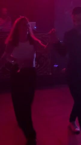 A woman in a white crop top and black pants dances with a man at a club. They are in front of a DJ booth with playing card decor, lit with red and green lights, moving to the music. Other people are visible in the background.