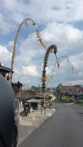 The video begins facing forward from the perspective of someone on a motorcycle. Ahead, a tall, curved bamboo pole decorated with greenery and colorful ornaments stands prominently on the right side of a paved road. The pole is a Penjor, a Balinese ceremonial offering. Behind it, two more Penjor poles are visible, one smaller and closer, and another further down the road. The road curves to the left, leading through a village. On the left side of the road, there are traditional Balinese houses with tiled roofs and open structures. Power lines crisscross the sky, which is partly cloudy with white cumulus clouds against a bright blue sky. The sun is shining, casting shadows. As the motorcycle moves forward, the Penjor poles and the village scenery pass by. The video ends with a wider view of the road and the village, with several Penjor poles visible along the roadside.