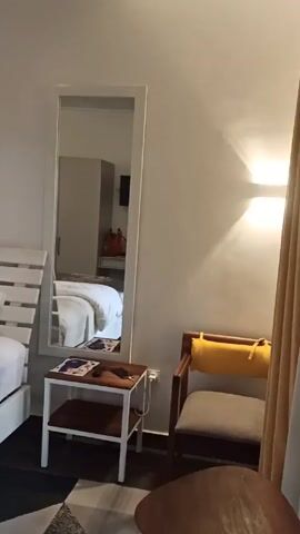 The video shows the interior of a bedroom. The room has white walls and a light-colored ceiling. A large bed with white linens and a gray throw is visible. The bed has a white, slatted headboard. There is a white framed mirror hanging on the wall beside the bed, reflecting a wardrobe. A small white table stands next to the mirror, holding a magazine and a remote control. A brown chair with a yellow cushion sits near the table. Two wall-mounted lights illuminate the area near the bed. A framed picture hangs above the bed.