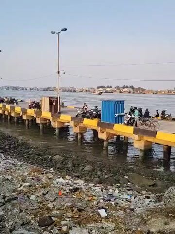The video shows a pier extending into the water. There are several motorcycles parked on the pier, and a few people are standing or sitting near them. One person is riding a motorcycle. There is a blue shipping container on the pier. The water is choppy, and there is a lot of debris on the shore in the foreground, including plastic bottles, wrappers, and other trash. In the background, there is a body of water with buildings and vegetation on the opposite shore. The sky is clear and the lighting suggests it is daytime, likely late afternoon or early evening.