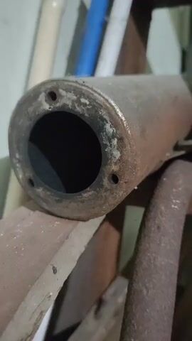 The video shows a close-up shot of a rusty, cylindrical object with a hole at one end. It appears to be a muffler or a part of an exhaust system. The object is resting on a wooden surface, and in the background, there are some vertical pipes or rods, one of which is blue. The lighting is dim, suggesting an indoor or shaded environment. The surface of the object is heavily corroded, with visible patches of rust and discoloration. The hole at the end is dark and appears to be hollow. The overall impression is that of an old, discarded mechanical part.
