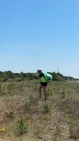 The video shows a person in a field picking up trash. The person is wearing a beige hat, a black shirt, black shorts, and a bright green safety vest. They are carrying a large green trash bag on their back. The field is mostly dry grasses with some green plants and yellow flowers scattered throughout. In the background, there are trees and shrubs along with what looks like a small white building or trailer. The sky is a clear blue with no clouds. The person uses a long handled grabber to pick up a post from the ground and adds it to the bag.