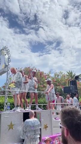 A group of men are dancing on a float. They are all wearing sparkly silver outfits. The float is decorated with stars and has speakers on it. The men are smiling and interacting with each other. The sky is blue with white clouds. There are trees and foliage in the background.