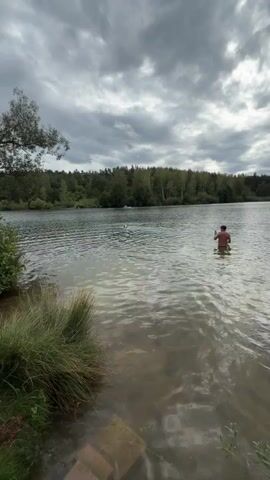 The video shows a scenic view of a lake on an overcast day. In the foreground, tall grass and some rocks are visible on the shore. The lake is calm with ripples on the surface. In the middle distance, a person is standing in the water, facing away from the camera, holding a phone. Further out on the lake, another person or object is visible in the water. The background consists of a dense forest of trees. The sky is cloudy, suggesting potential for rain.