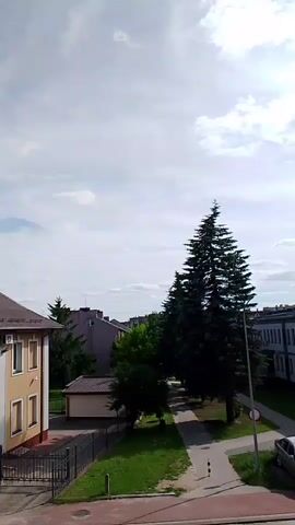 A short video clip pans across a street view with a building on the right, a large evergreen tree in the center, and a building on the left. Several cars are parked along the street in front of the building on the right. Some people are visible walking in front of the building. The sky is partly cloudy and the scene appears to be outdoors during the daytime.