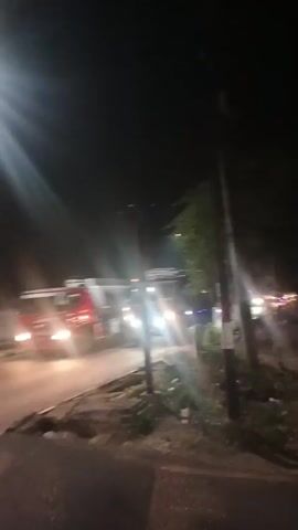 The video begins with a dark outdoor scene at night, illuminated by bright lights, likely from vehicles. A large red truck with its headlights on is visible, followed by a portion of another vehicle in the foreground on the left. The camera then pans to show a black helmet and the back of a person wearing a red jacket. The view shifts to the perspective of someone riding a motorcycle at night, with the road ahead visible. Several other motorcycles are seen passing by, some blurred due to motion. A street with a curb, vegetation, and a utility pole is visible on the right. Further ahead, there appears to be a sign for a village or area. The video ends with continued motorcycle traffic on the dark road.