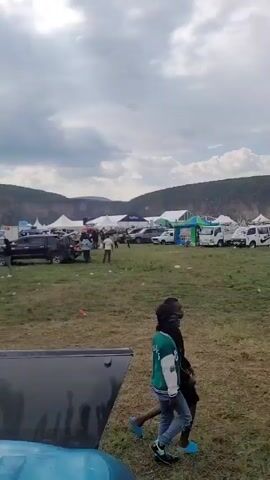 The video shows a wide outdoor scene under a partly cloudy sky. The ground is grassy, and there are several people, mainly children, walking through the area. In the background, there is a line of vehicles, tents, and other structures, suggesting some sort of event or gathering. The backdrop features a low-lying mountain range.