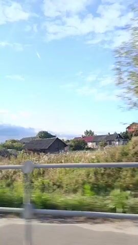 A video taken from a moving vehicle shows a rural landscape under a bright blue sky with scattered clouds. The view includes a variety of houses, some with modern architecture and others appearing older with dark roofs. There are fences, some made of corrugated metal and others of metal rails, bordering grassy areas and fields of tall dry plants. Trees are visible behind the houses and fences. The overall impression is a serene, sunny day in a village setting.