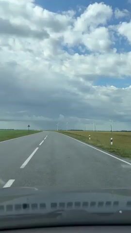 The video shows a view from a vehicle traveling on a paved road through a rural landscape. The road has white dashed lines indicating lanes. To the right of the road is a field of tall, golden grain, likely wheat or barley. In the distance, several white wind turbines are visible, standing tall against a sky filled with a mix of dark storm clouds and lighter, puffy white clouds. The sun is not directly visible, but there is ambient light suggesting it is daytime. The overall impression is of a vast, open agricultural area under a dynamic, cloudy sky. There are no people or other vehicles clearly visible in the immediate vicinity. The road curves slightly to the left in the distance.