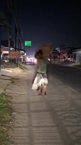 At night, a man walks along a paved sidewalk next to a road, carrying several rectangular cardboard boxes on his shoulders. He is dressed in a striped shirt and patterned sarong, walking barefoot. The road has streetlights and car headlights are visible in the distance. There are buildings on either side of the road and a street sign is visible on the sidewalk next to the man.