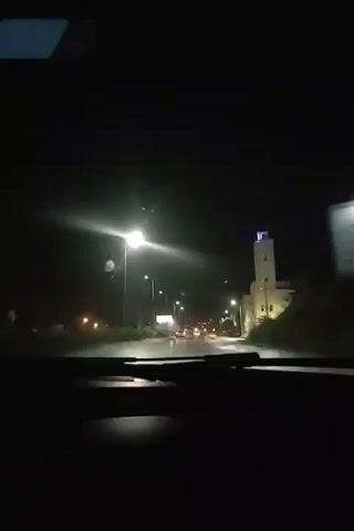 The video shows a nighttime view from inside a car looking forward through the windshield. Streetlights illuminate a wet road with a few cars driving in the distance. To the right, a building with a tower and a blue light on top is visible. The dash of the car is in the foreground, and the windshield wipers are visible at the bottom of the frame. The weather appears to be rainy.