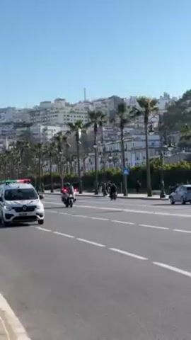 The video depicts a motorcade moving down a road in a city on a sunny day. The motorcade includes a white police car with green and red lights on top, followed by police motorcycles. Behind the motorcade, there's a black van and other civilian cars. Pedestrians are walking on the sidewalk alongside the road. The city buildings are visible in the background, with a mix of white and other colored buildings, palm trees, and street lights lining the road.