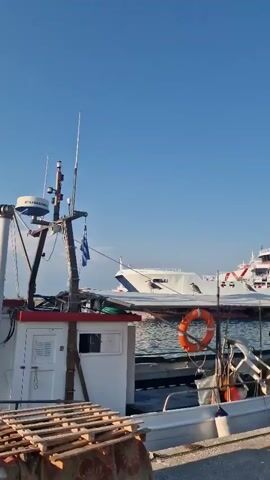 The video shows a harbor scene. In the foreground, there is a small fishing boat with a white cabin and a red trim. A life preserver, an orange ring, is attached to the side of the boat. Wooden pallets are stacked on the left side of the boat. In the background, a large white ferry with blue and red stripes is docked in the water. The sky is clear and blue, indicating good weather. The scene appears to be peaceful and calm.