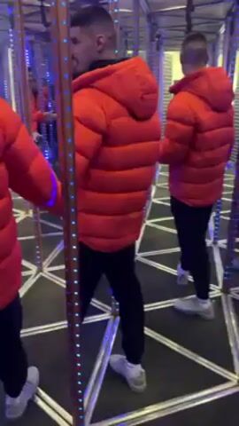 A man in a bright red puffy jacket, black pants, and white sneakers is walking inside a mirror maze. The maze's floor has a pattern of white triangles with black edges. The mirrors are framed by vertical strips with blue LED lights. The man appears to be feeling his way through the maze, using his hand to check for solid surfaces. His reflections appear multiple times in the surrounding mirrors, creating a disorienting effect.