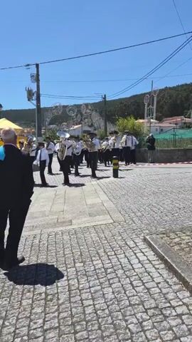 A marching band is parading on a cobblestone street in a small town. Most band members are wearing white shirts, black pants, black shoes, and black caps with white trim. Some wear green ties. They are playing various instruments, including trombones, saxophones, tubas, and drums. The band is marching towards the camera, and a man in a dark suit is standing on the left side of the frame, watching them. In the background are houses, a hill covered in trees, and decorative lights. The sky is clear and blue.