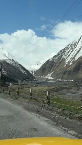 From a vehicle's perspective, a scenic view of a valley unfolds. Snow-capped mountains rise on both sides, their peaks gleaming under a partly cloudy, blue sky. A river snakes through the valley floor, alongside patches of green fields. A weathered, grey road stretches beside a simple fence, while power lines run overhead, supported by wooden poles. The dashboard of the vehicle, visible at the bottom of the frame, is bright yellow.