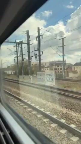The video shows a view from inside a moving train, looking out the window at the passing scenery. The train tracks are visible in the foreground, running parallel to the train. Beyond the tracks, there are buildings, trees, and utility poles. The sky is partially cloudy. As the train moves, it passes by a station platform with a few people waiting. The train appears to be moving at a moderate speed.
