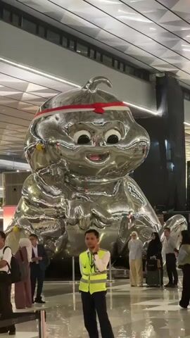 The video shows a large, shiny, silver inflatable sculpture of a character with large eyes and a smiling mouth, wearing a red and white headband. The sculpture is in a large, modern indoor space with a high, patterned ceiling and bright lighting. Several people are visible around the sculpture. In the foreground, a person wearing a bright yellow safety vest and dark pants stands looking towards the sculpture. To the left of the sculpture, a person with a backpack stands and looks away from the camera. To the right of the sculpture, several people, some wearing headscarves, are gathered, with one person pulling a suitcase. The overall environment appears to be a public space, possibly an airport or a large exhibition hall.