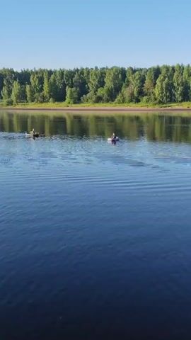 Two people are on a river in small boats. The person on the left is in a dark boat and appears to be paddling. The person on the right is in a lighter colored boat and is also paddling. They are both moving away from the camera. The river is wide and calm, with ripples on the surface. A dense line of green trees forms the background, with a clear blue sky above. The sun is shining, casting reflections on the water.