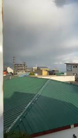 The video presents a series of rooftop views under changing sky conditions. Initially, a green corrugated iron roof is visible under a cloudy sky. The video then transitions to show other rooftops, mostly of corrugated iron with varying degrees of wear and rust, set against skies transitioning from partly cloudy to sunny. Buildings and vegetation are visible in the background. The camera pans slightly to show more of the surrounding urban environment.