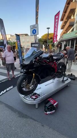 The video shows a black motorcycle on display outdoors on a metal stand. The motorcycle is a sportbike with a red frame and gold wheels. A red and black helmet sits on the ground in front of the motorcycle. In the background, there are people, buildings, and signs. One sign reads "DUCATI." The setting appears to be a street or public square, possibly during an event. The overall lighting suggests it is either early morning or late afternoon.