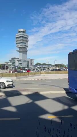 This media file captures a view of an airport terminal on a sunny day with a bright blue sky and scattered white clouds. The main subject is a modern airport building with a distinctive facade featuring diamond-shaped reflective panels and a vertical green wall. The building has a contemporary architectural style with a covered entrance area.

In the foreground, a blue bus is parked, suggesting passenger transport or shuttle services. The bus has the text "GONECTO" and "HYBRID" visible on its rear. There are also signs indicating directions and airport services. One prominent sign reads "Uloz Entrance" and shows arrows pointing to "Terminal 2," "Registracija 101-319," "Terminal 1," and "Agency Offices 401-608," as well as "Bus Public Transport." The Cyrillic text "БЕОГРАД" is prominently displayed on the building, indicating the location is Belgrade Airport in Serbia.

Several people are visible near the entrance, some with luggage, suggesting travelers arriving or departing. The overall scene depicts a typical airport environment, busy with activity related to air travel. The lighting suggests it is daytime, likely late morning or early afternoon, given the bright sunlight casting distinct shadows. The weather appears pleasant, with clear skies contributing to a bright and open atmosphere.