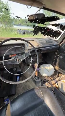 This media file showcases a severely dilapidated, older model car, likely a Soviet-era Moskvich, in a state of disrepair. The interior is visible, revealing a worn steering wheel with a "BOSS" sticker, a gear shift, a plastic jug, and various other items strewn across the floor and seats. The car's exterior shows significant rust, peeling paint, and a damaged windshield. A broken rear window is also evident, with a piece of glass precariously leaning inside.

The scene appears to be outdoors, possibly a parking lot or roadside, under a clear, sunny sky, suggesting it is daytime. In the background, a store with a prominent sign reading "МАГНИТ КОСМЕТИК" (Magnit Kosmetik) is visible, indicating the car is located in Russia, specifically in a place called Solnechny. The overall impression is one of neglect and abandonment, with the car seemingly left to decay. No people are present in the media.