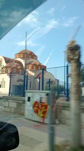 The video shows a side view of a church with a dome. The church has arched windows and doors. There are trees in front of the church. The sky is blue and there are some clouds. The video is shaky, suggesting it was filmed from a moving vehicle. The overall scene appears to be in a sunny, possibly Mediterranean, location.