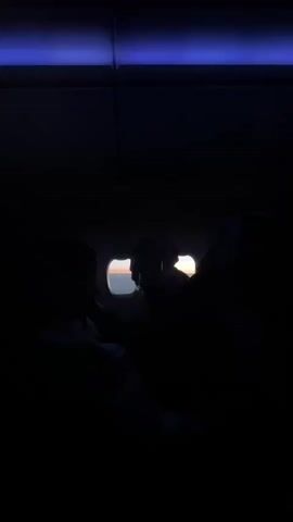 The video shows the interior of an airplane at dusk or dawn. The cabin is dark, with the window providing the only significant light source. Passengers are silhouetted against the light from the window, where the sky is visible. The horizon is distinct, with the sun illuminating a small section of the sky. The view is mostly still, focusing on the window and the surrounding area.