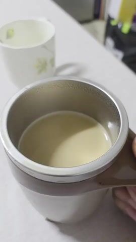 A close-up video shows a person's hand holding a mug with a metal interior, as liquid inside is being stirred by a spinning mechanism. The liquid is a light beige color and swirls vigorously, creating a vortex in the center. Another mug, white with a floral pattern, is visible in the background. The surface the mug is resting on is light-colored and appears to be a table or countertop. The lighting is indoor and appears to be natural light from a window.